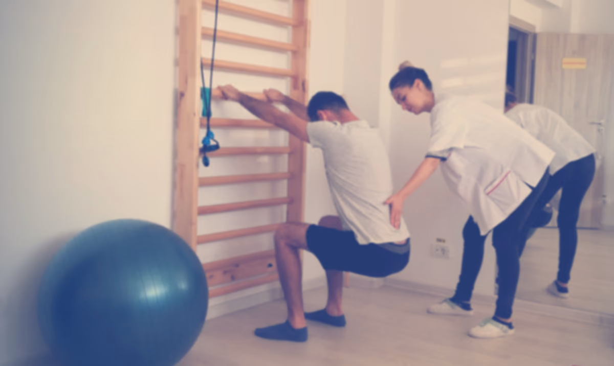 Kinetoterapeut care ghidează exercițiul unui pacient la spalier, într-o sală de recuperare medicală